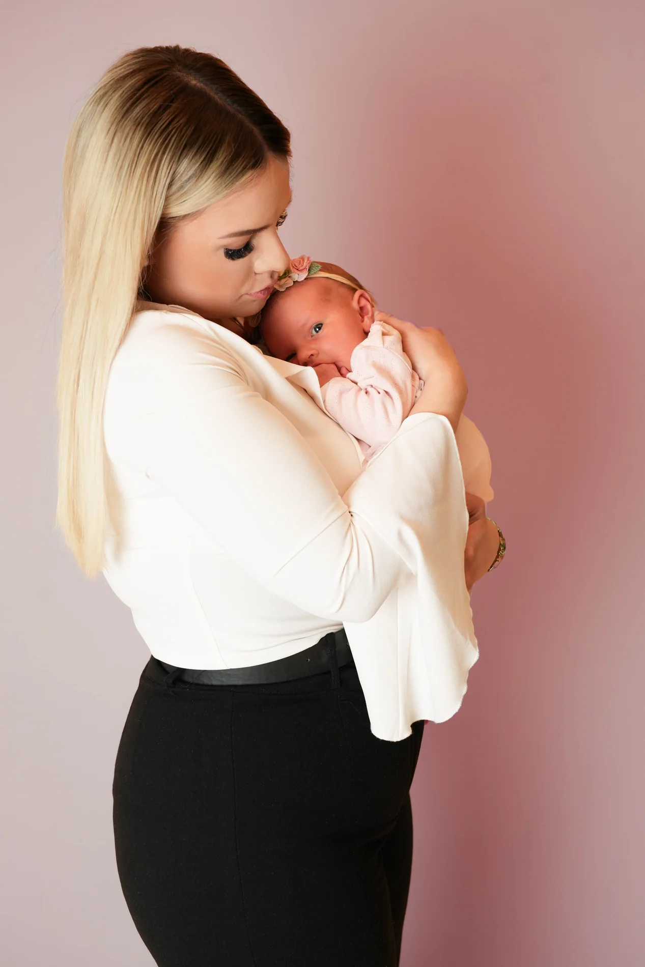 Frau im Seitenprofil küsst ein Baby mit Blumenstirnband vor einer rosa Wand