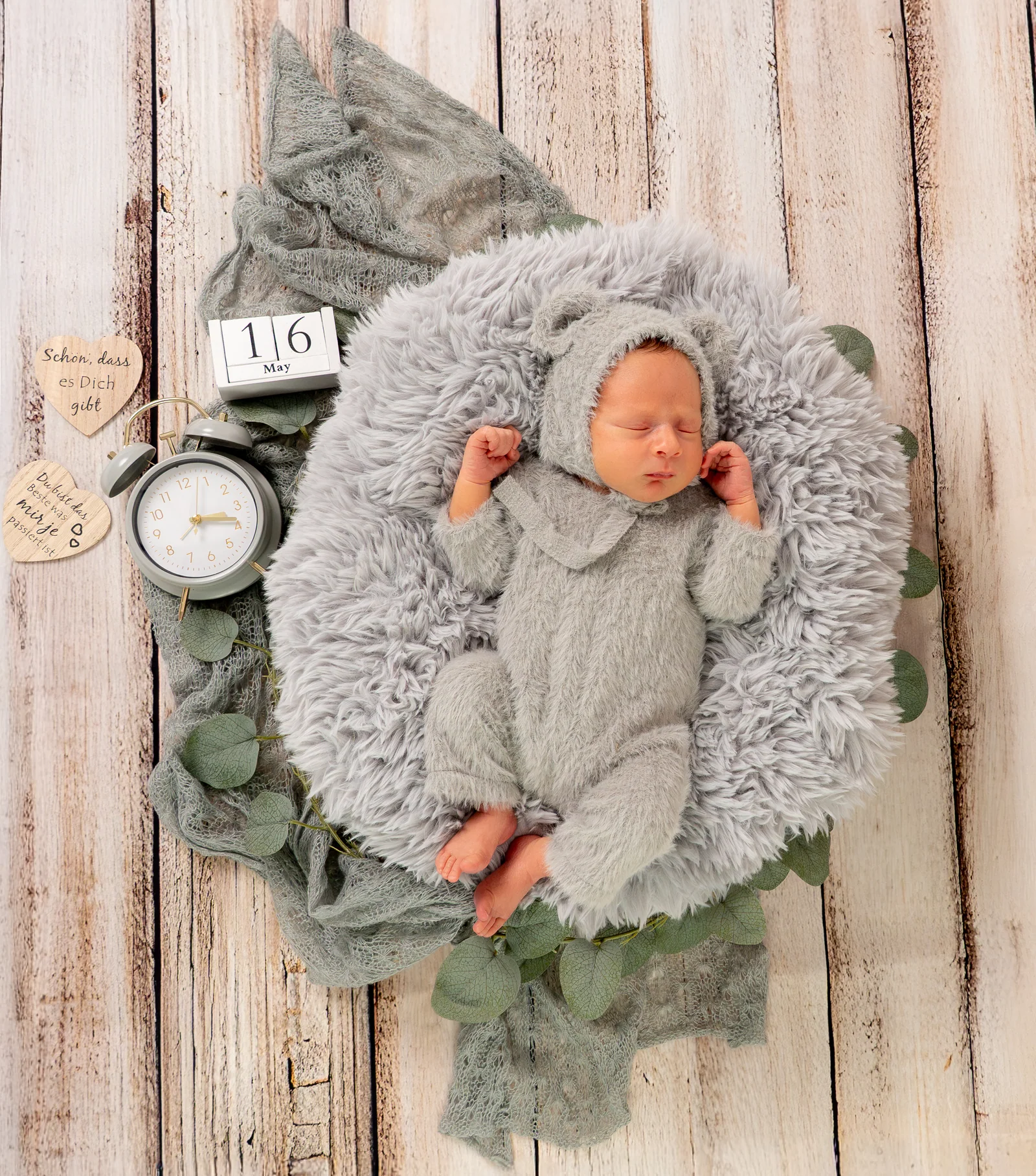 Draufsicht auf ein Baby im grauen Bärenanzug in einem flauschigen Körbchen mit Blättern und Wecker