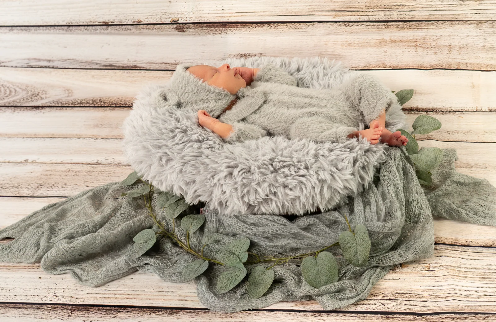 Baby im grauen Strickanzug liegt mit ausgebreiteten Armen im Körbchen vor hellen Holzbrettern