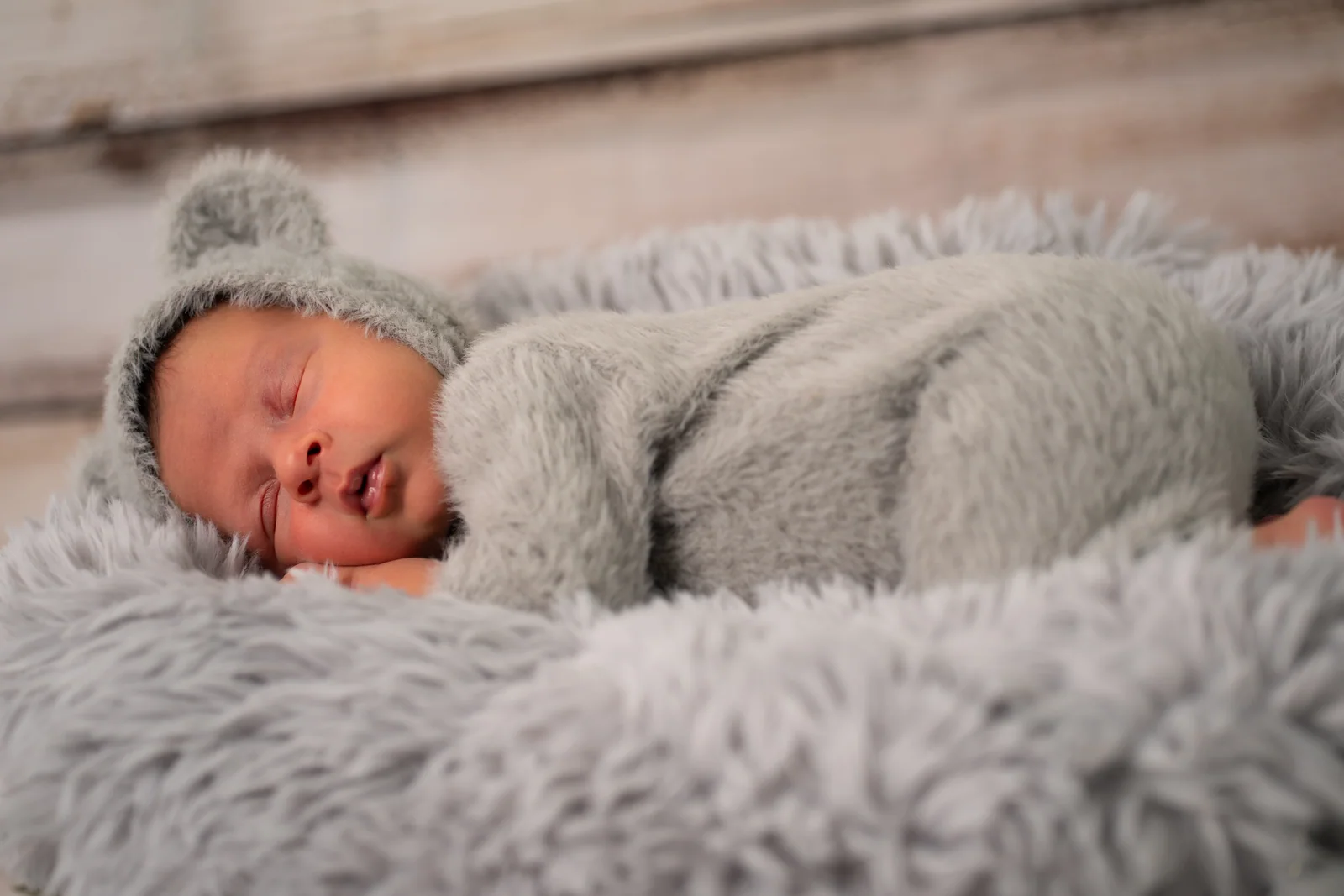 Baby im flauschigen grauen Anzug schläft eingerollt auf der Seite vor einer Holzwand