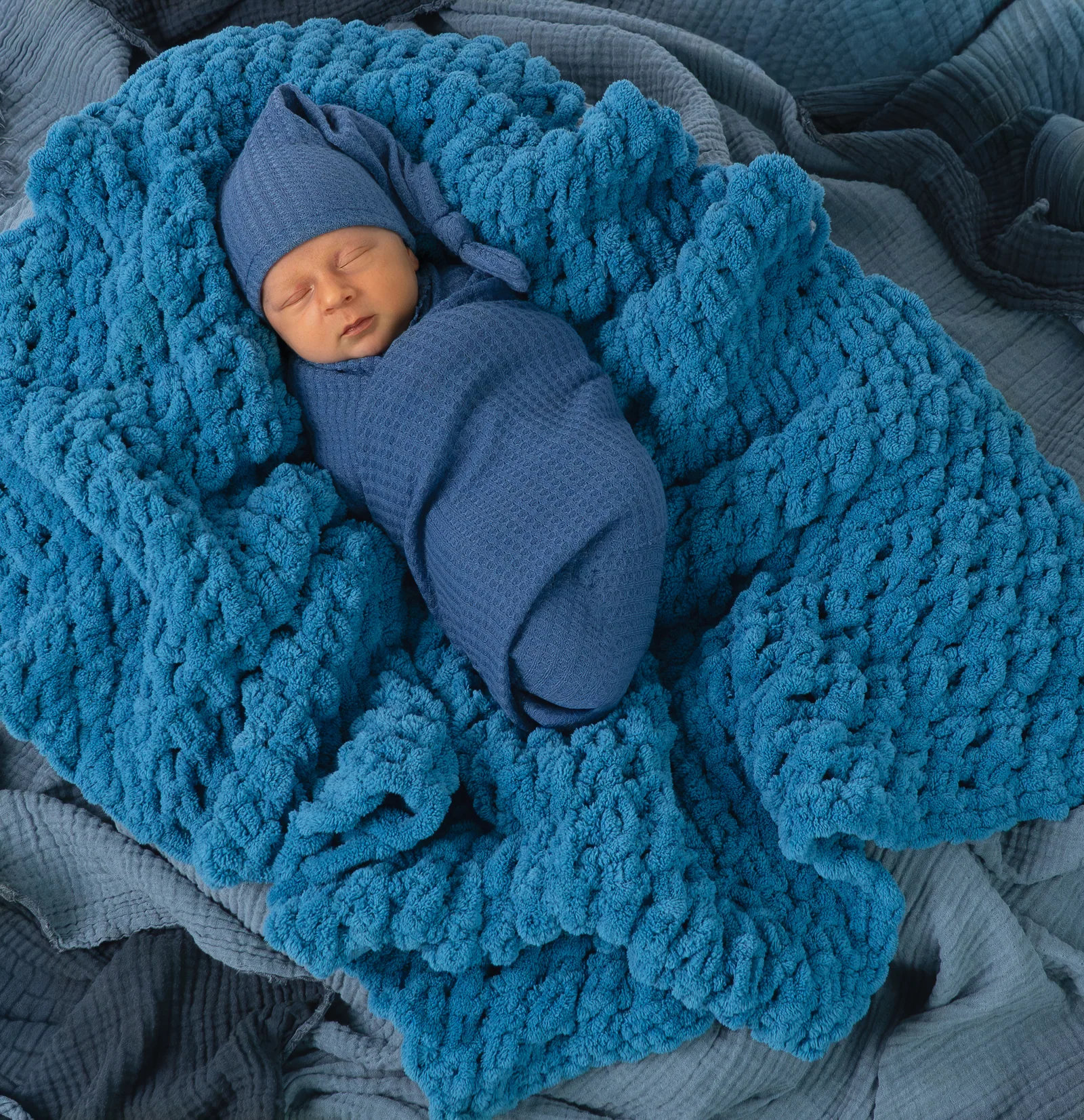 Von oben fotografiertes Baby in blauem Wickeltuch auf einer türkisblauen groben Strickdecke