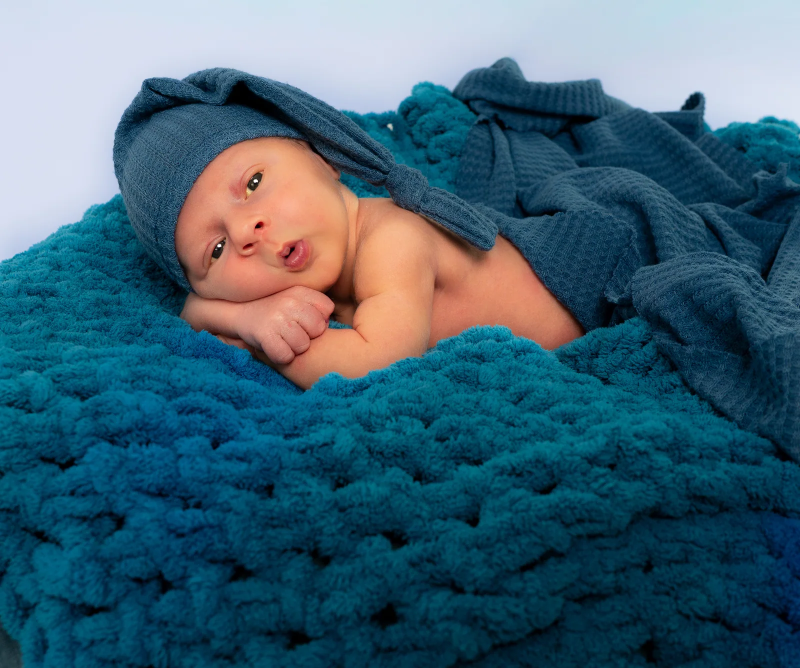 Waches Baby mit blauer Zipfelmütze legt den Kopf auf seine Hände und schaut in die Kamera