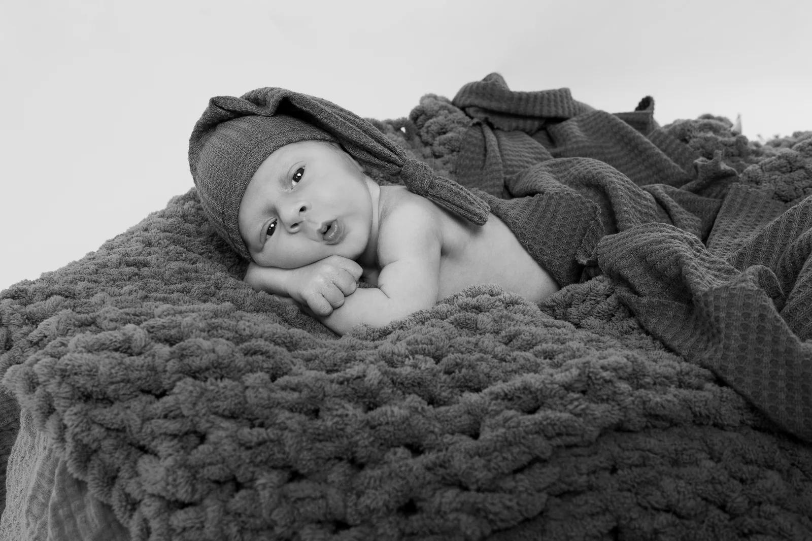 Schwarzweißporträt eines wachen Babys mit Zipfelmütze, das auf einer groben Strickdecke liegt