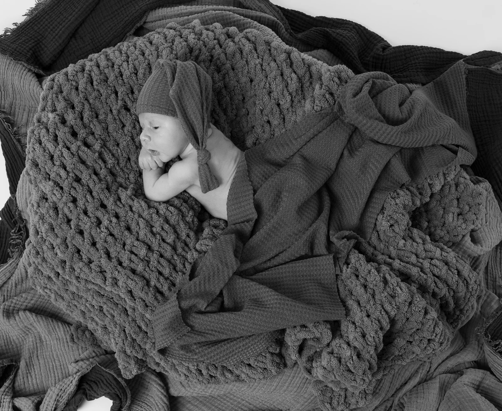 Schwarzweiße Draufsicht auf ein Baby mit langer Strickmütze, umgeben von locker gefalteten Decken