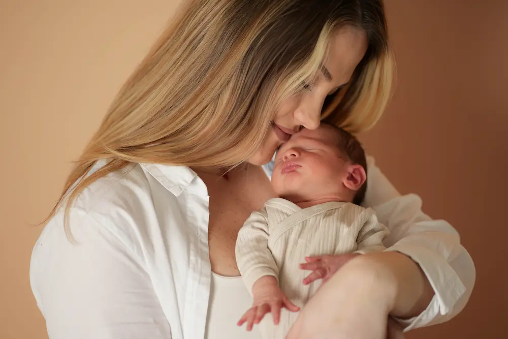 Mutter in weißer Bluse küsst ihr schlafendes Baby auf die Stirn