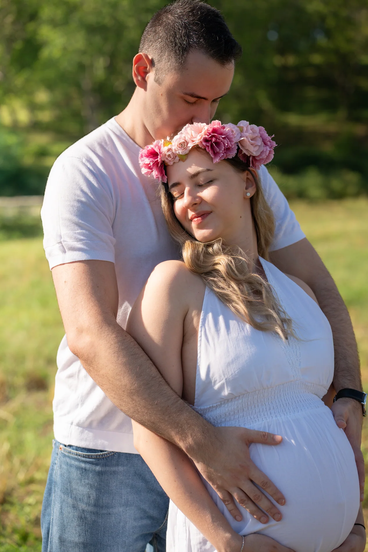 Ein Mann küsst eine schwangere Frau mit Blumenkranz im Haar unter grünen Bäumen