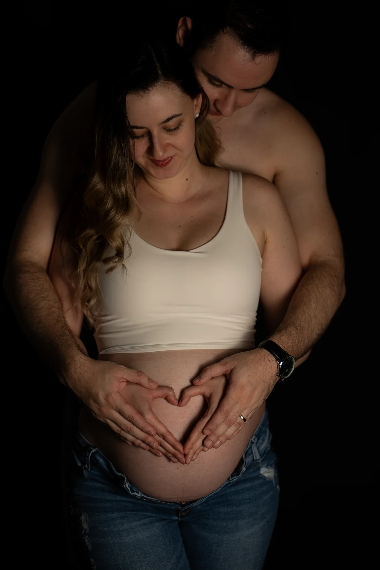Paar vor schwarzem Hintergrund mit herzförmig auf dem Babybauch gelegten Händen
