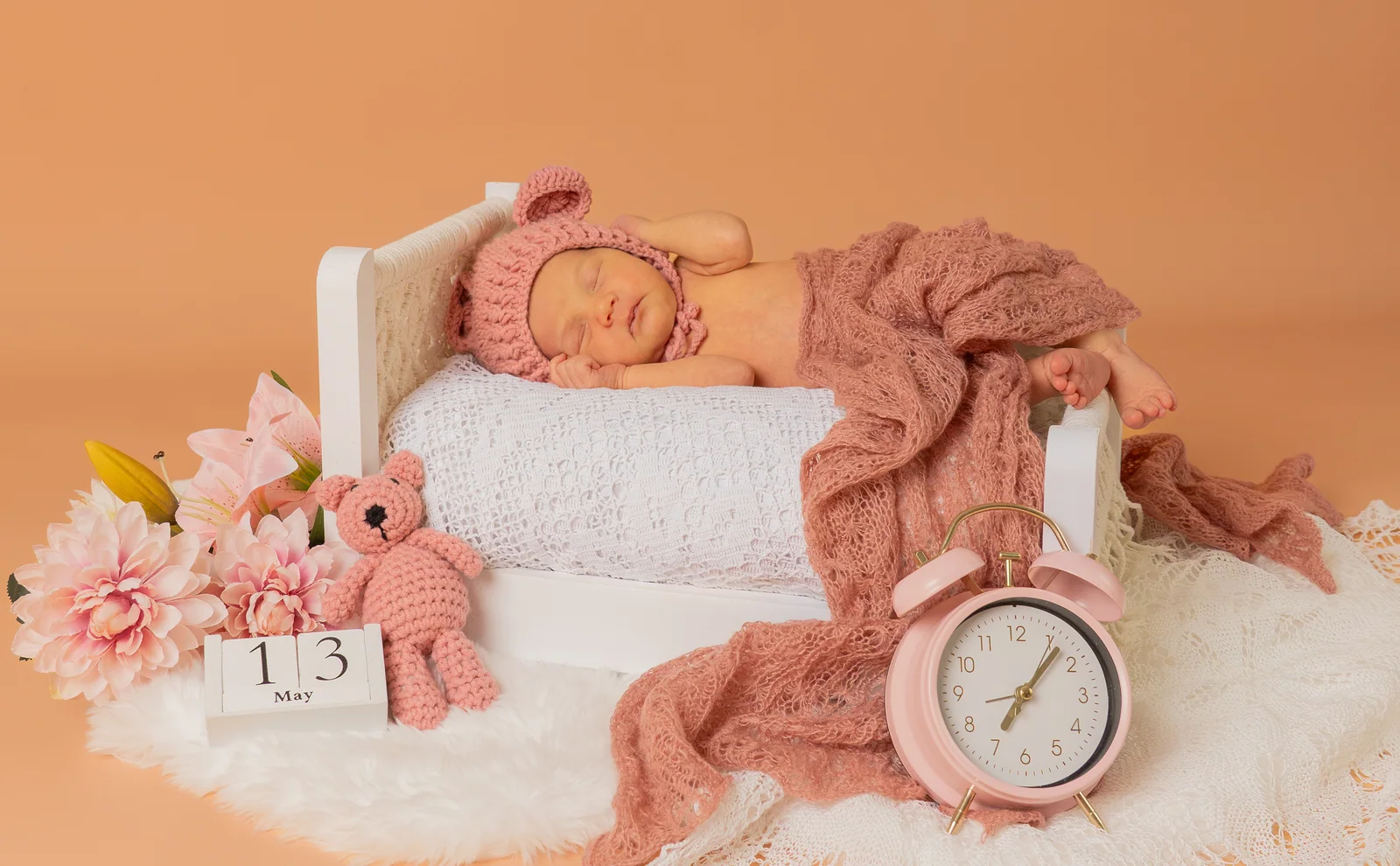 Baby mit rosa Mütze im kleinen Bett, daneben Blumen, Strickhase und Wecker